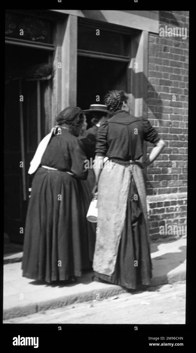 Drei Frauen in einer Londoner Straße, die auf dem Bürgersteig vor einem Haus mit Terrasse stehen. Einer trägt eine lange Schürze und einen Krug. Stockfoto