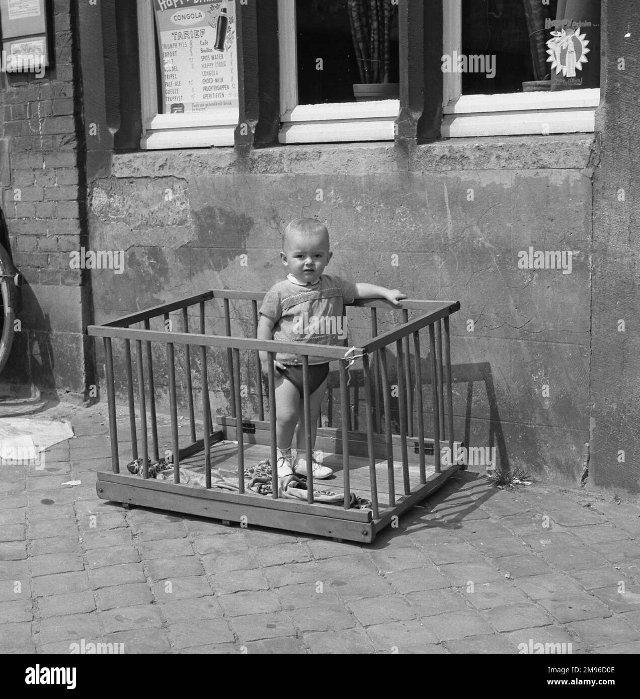 Ein kleiner Junge steht in einem Laufstall auf einem Gehweg vor einem Café. Stockfoto