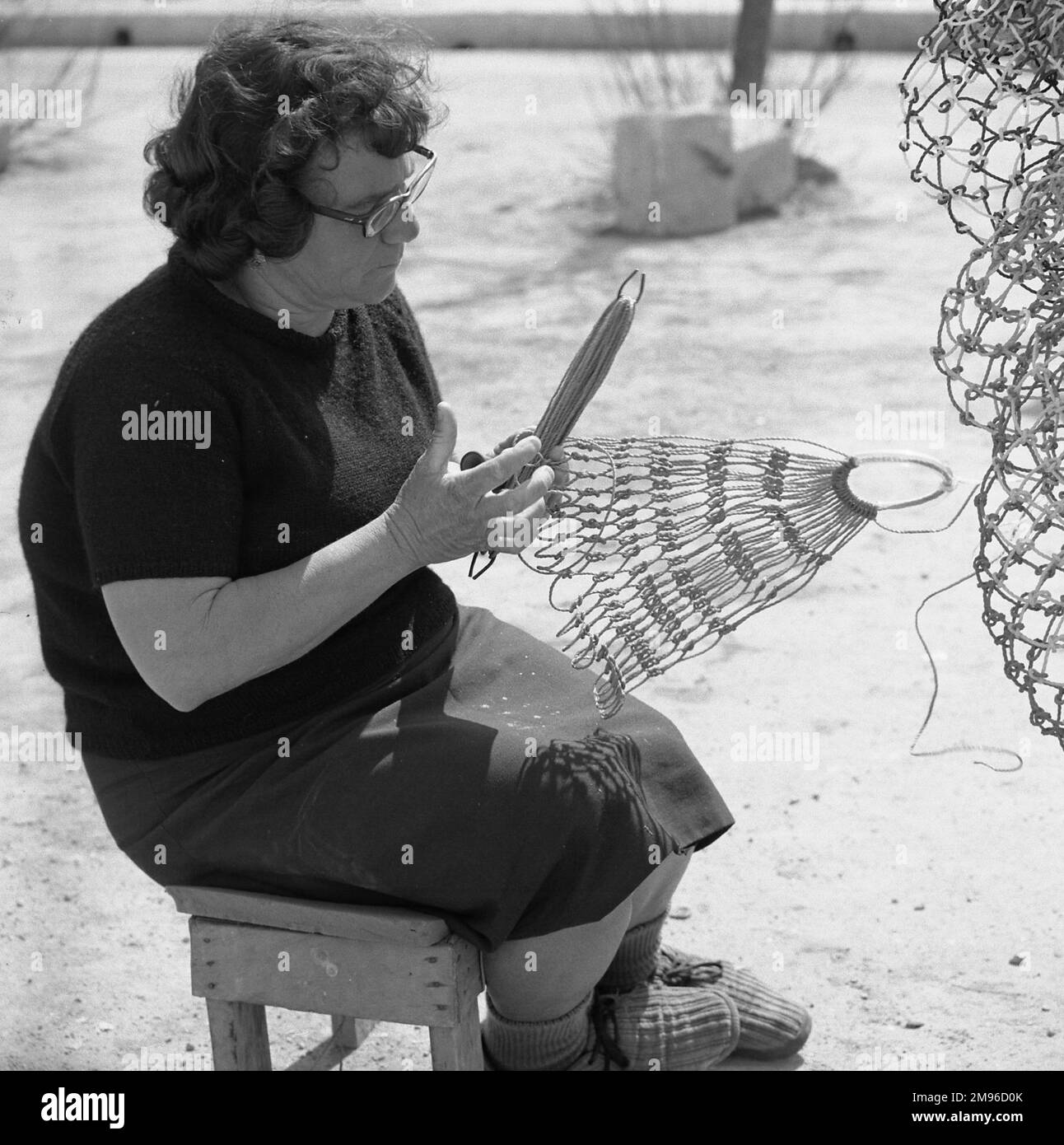 Eine Frau mittleren Alters, die in der Sonne sitzt und ein Fischernetz herstellt oder repariert. Stockfoto