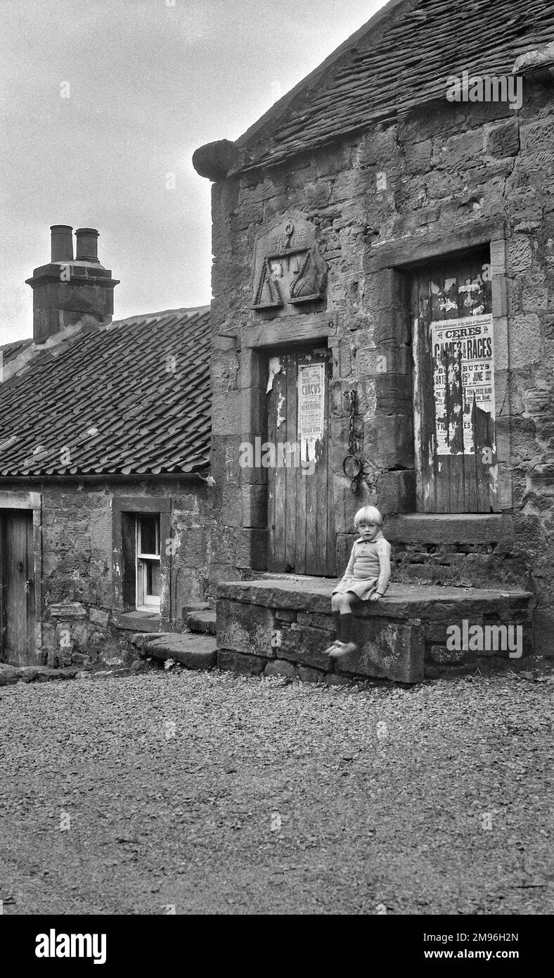 Straßenszene in den schottischen Highlands, mit einem kleinen Mädchen, das auf einer Steintreppe sitzt. Eine Steinschnitzerei einer Waage trägt die Inschrift: Gott segne die Gerechten. Stockfoto