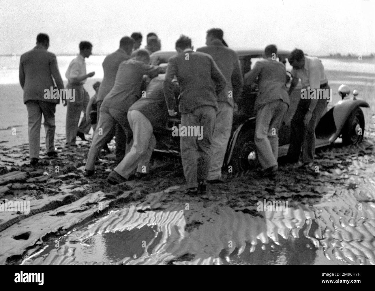 Eine Gruppe von Männern, die ein Auto auf einen nassen Sandstrand schieben. Stockfoto