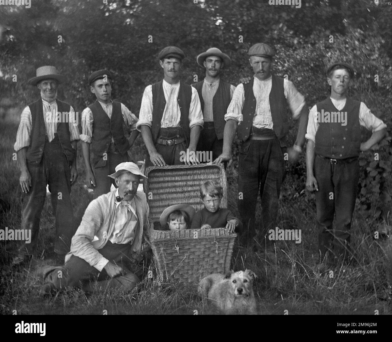 Sieben Männer, zwei Kinder, die in einem Korb sitzen, und ein kleiner Terrier-Hund, auf einem Feld. Stockfoto
