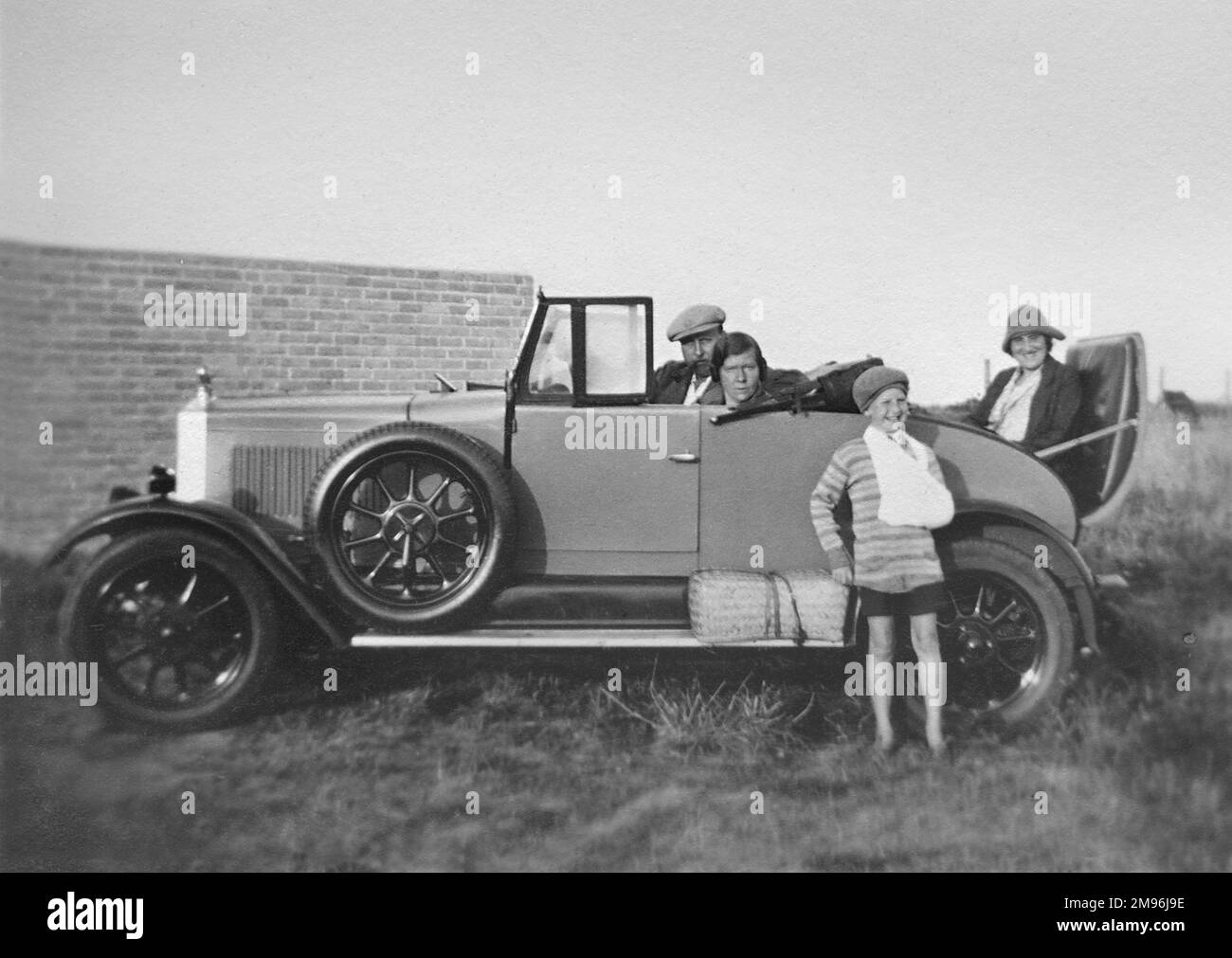 Drei Personen sitzen in einem offenen Auto. Ein lächelnder Junge steht vorne mit seinem Arm in einer Schlinge. Stockfoto