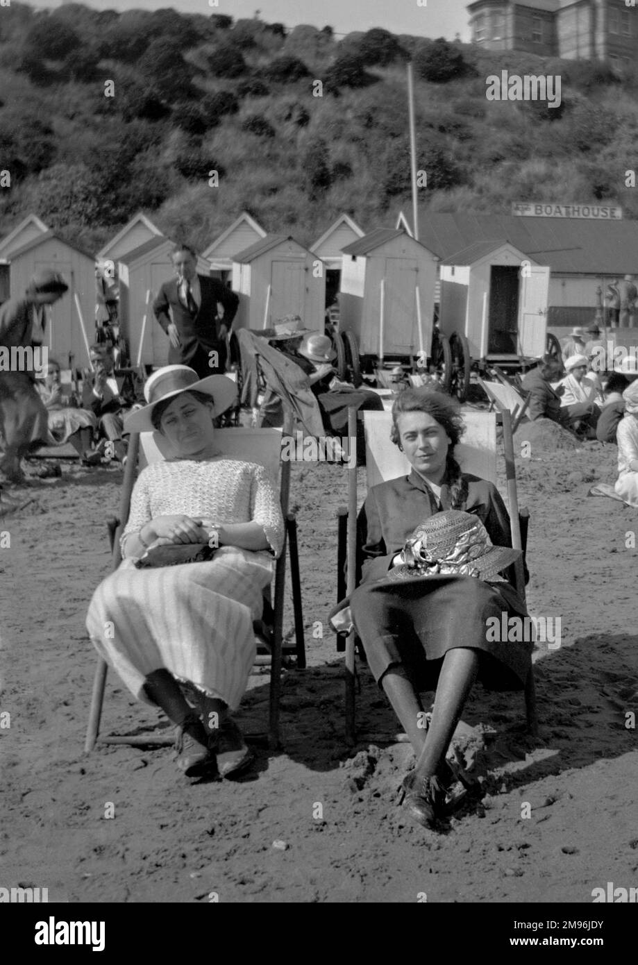 Zwei junge Frauen sitzen am Strand in Liegestühlen, mit Badehütten im Hintergrund. Stockfoto