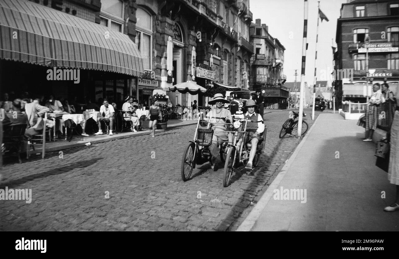 Eine Frau und ein Junge auf einem vierrädrigen Zweisitzer-Fahrrad (auch bekannt als Velo-Doppelzimmer) in Brügge, Belgien. Die Leute sitzen an Tischen vor einem Café auf der linken Seite. Stockfoto