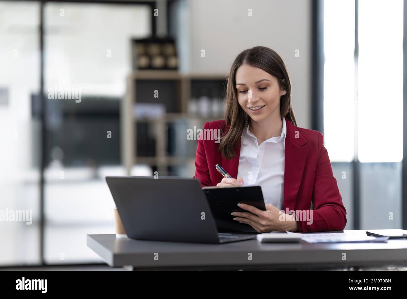 Lächelnde Frau, die am Schreibtisch sitzt und an einem Laptop arbeitet, Notizen in einem Notebook macht, glückliche Millennials, die online arbeitet, sich ein Webinar mit dem Computer ansieht und Stockfoto