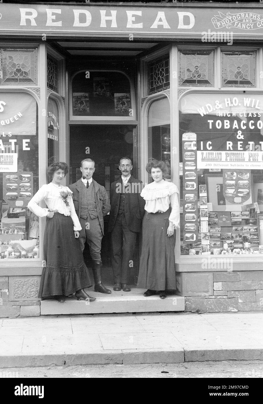 Redhead's Shop, Coniston, befindet sich in der Yewdale Road und wird immer noch als Zeitungskiosk/Souvenirladen geführt. Die Männer in der Tür sind die Freunde des Fotografen Wilson und Nelstrop, und die Frauen werden als "Annie und Kate" identifiziert, die dort gearbeitet haben. Stockfoto