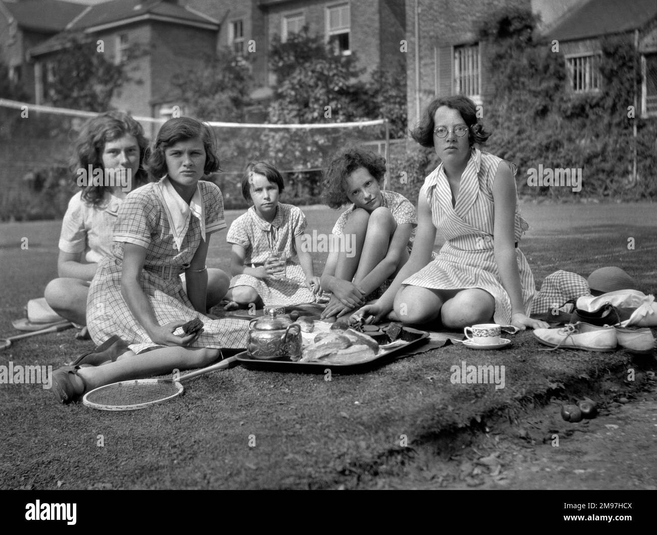 Fünf Mädchen mit einem Teetablett im Garten an einem Sommertag. Auf dem Rasen im Hintergrund wird ein Badmintonnetz aufgestellt. Stockfoto