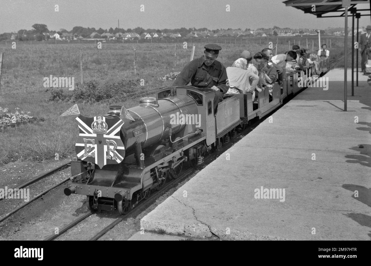 Erwachsene und Kinder, die in einer Miniatur-Dampfeisenbahn mit einer britischen Flagge vorne fahren. Stockfoto