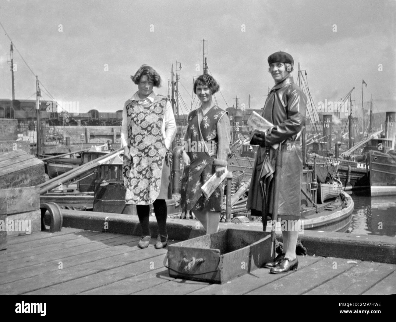 Drei Frauen stehen am Hafen, zwei tragen Schürzen, die dritte trägt einen Ledermantel. Stockfoto