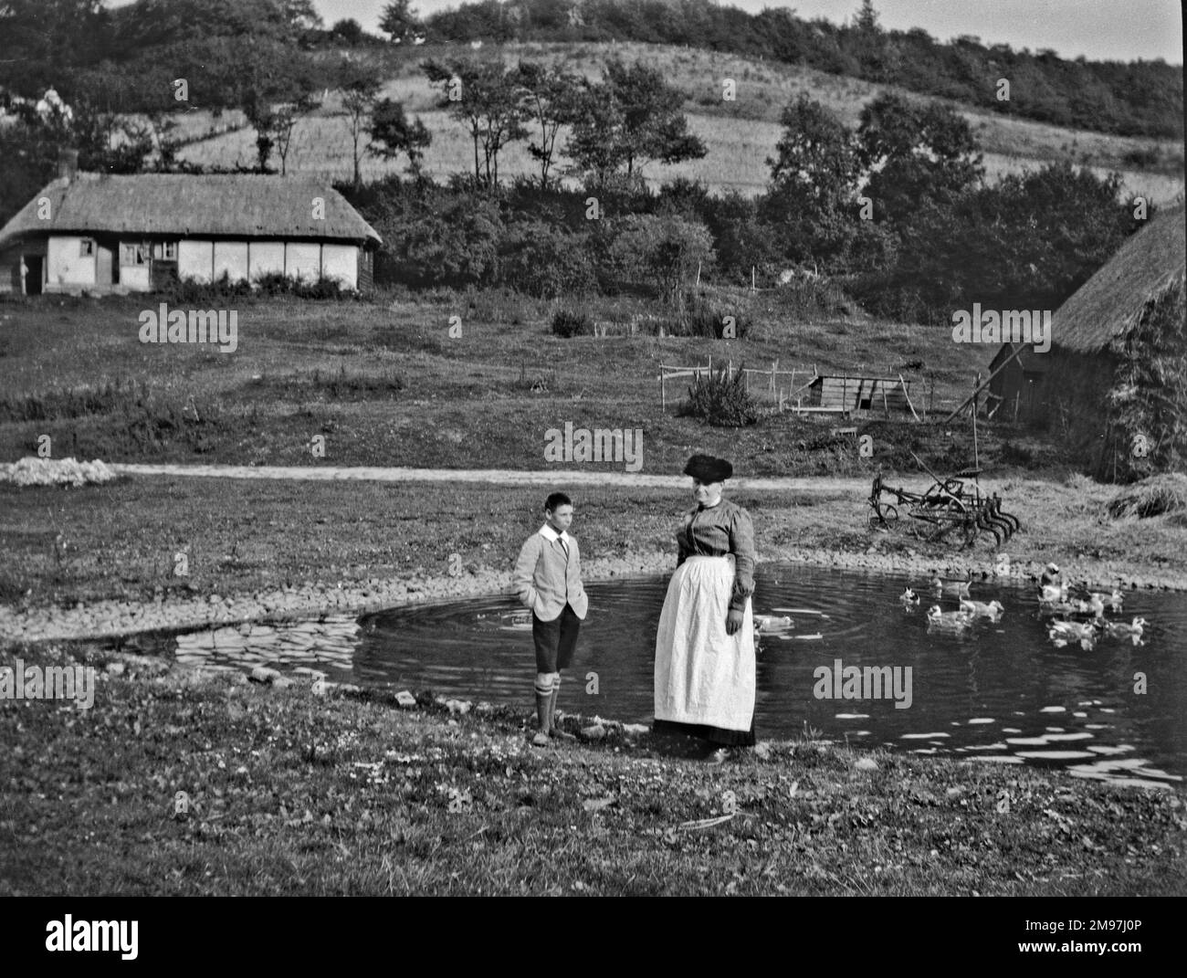Frau und Junge stehen an einem Ententeich in der Nähe einer strohgedeckten Hütte. Stockfoto