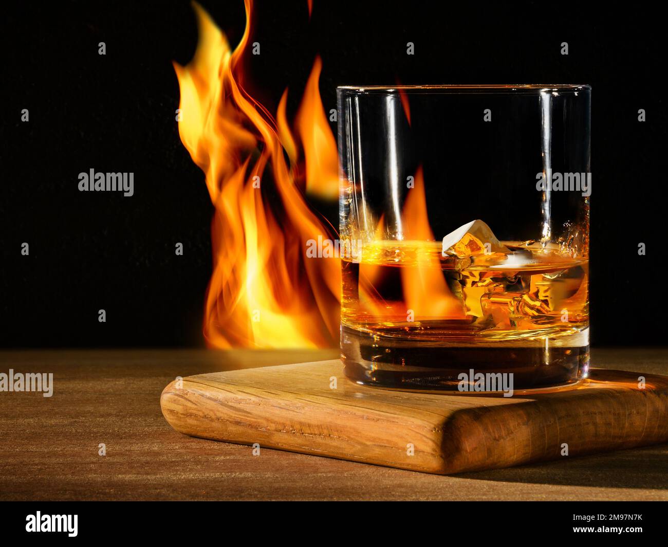 Glas mit Whiskey mit Eis auf Holztisch und schwarzem Hintergrund mit Feuer Stockfoto