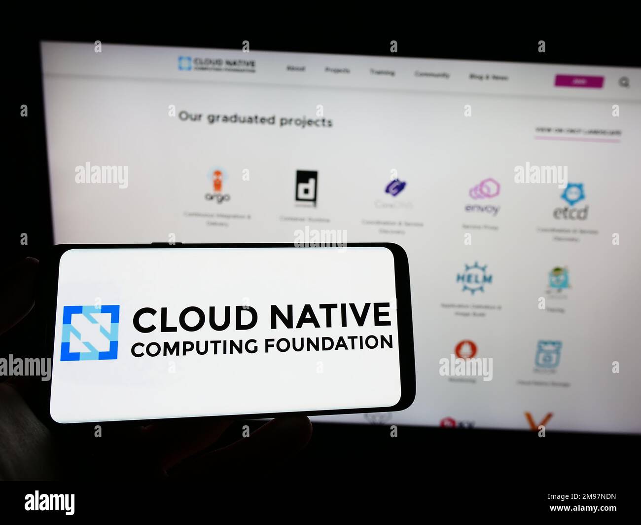 Person, die ein Mobiltelefon mit dem Logo der Cloud Native Computing Foundation (CNCF) auf dem Bildschirm vor der Webseite hält. Konzentrieren Sie sich auf das Display des Telefons. Stockfoto