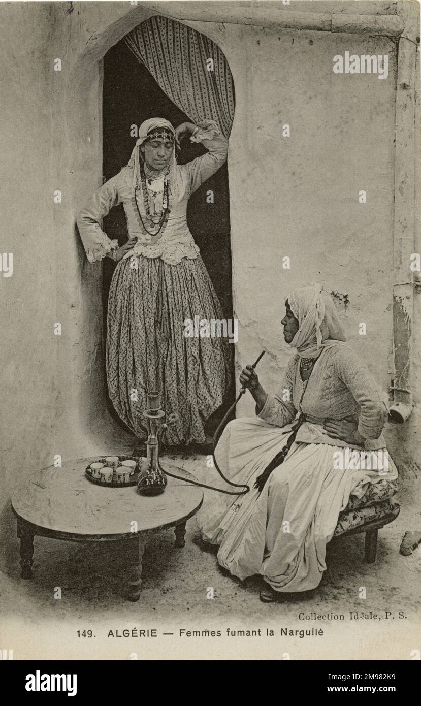 Zwei Frauen rauchen aus einer Wasserpfeife in Algerien, Nordafrika. Eine Frau sitzt und hält den Stiel der Sheesha-Pfeife nahe ihrem Mund, bereit zum Einatmen. Die andere Frau steht in einer Tür in ihrer Nähe, beobachtet und wartet darauf, dass sie an der Reihe ist. Beide Frauen zeigen traditionelle Kleidung und Kopftücher, die für die Region typisch sind. Stockfoto