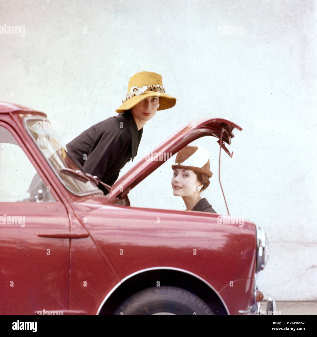 Tatler-Cover-Fotoshooting mit Fokus auf Hüte und die Motor Show -- zwei weibliche Models, die sich mit einem roten Mini-Auto posieren. Für das Cover der Tatler-Ausgabe vom 21. Oktober 1959 wurde eine Variantenaufnahme dieses speziellen Fotoshooting verwendet (siehe Abbildung 11064837). Stockfoto