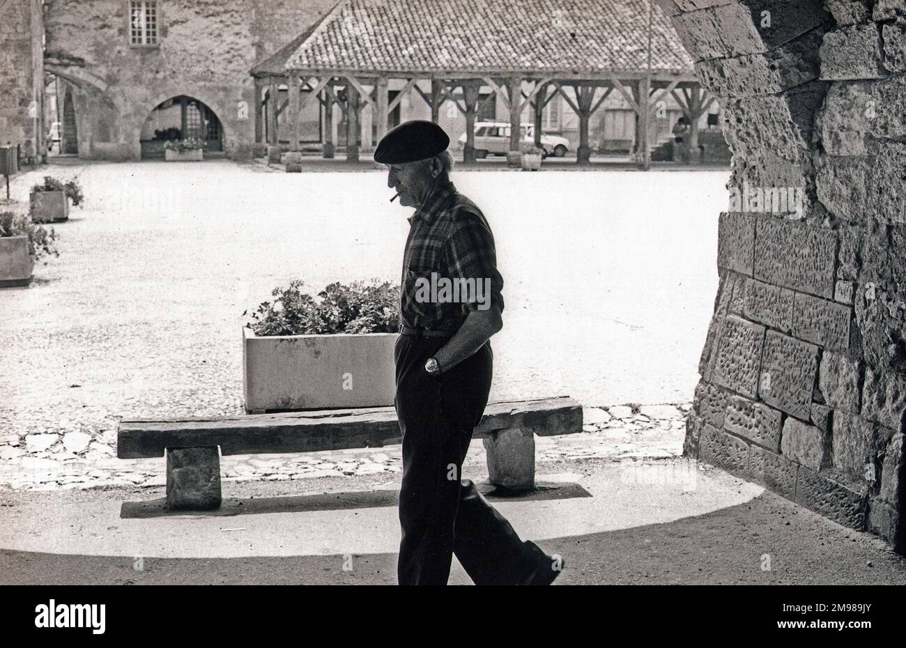 Ein Mann mit Baskenmütze, der raucht, während er an einem sonnigen Marktplatz in Frankreich vorbeigeht. Stockfoto