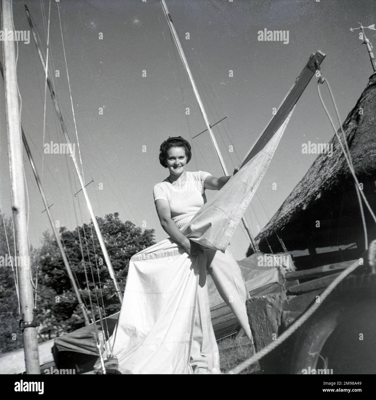 Barbara Hepworth (1903-1975), englische Künstlerin und Bildhauerin, die das Segel einer Yacht hält, mit einem strohgedeckten Häuschen auf der rechten Seite. Stockfoto