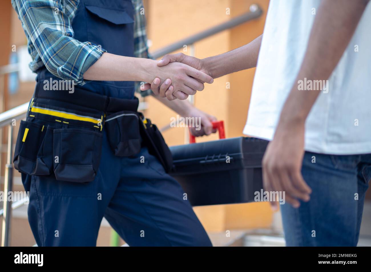 Freigestelltes Foto eines Reparaturwerkers, der einen Werkzeugkasten hält, während er mit seinem Kunden im Freien die Hand schüttelt Stockfoto