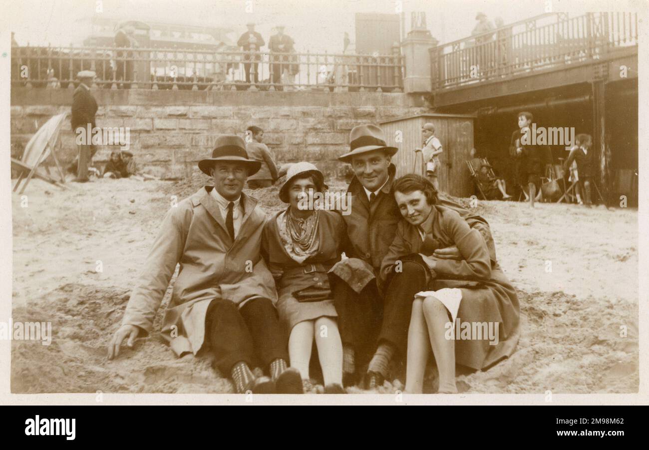 Typische britische Sommerszene - zwei Paare, die sich am Strand warm einwickeln und ihn mit Mänteln und Hüten aushöhlen. Vielleicht in Blackpool aufgenommen. Stockfoto