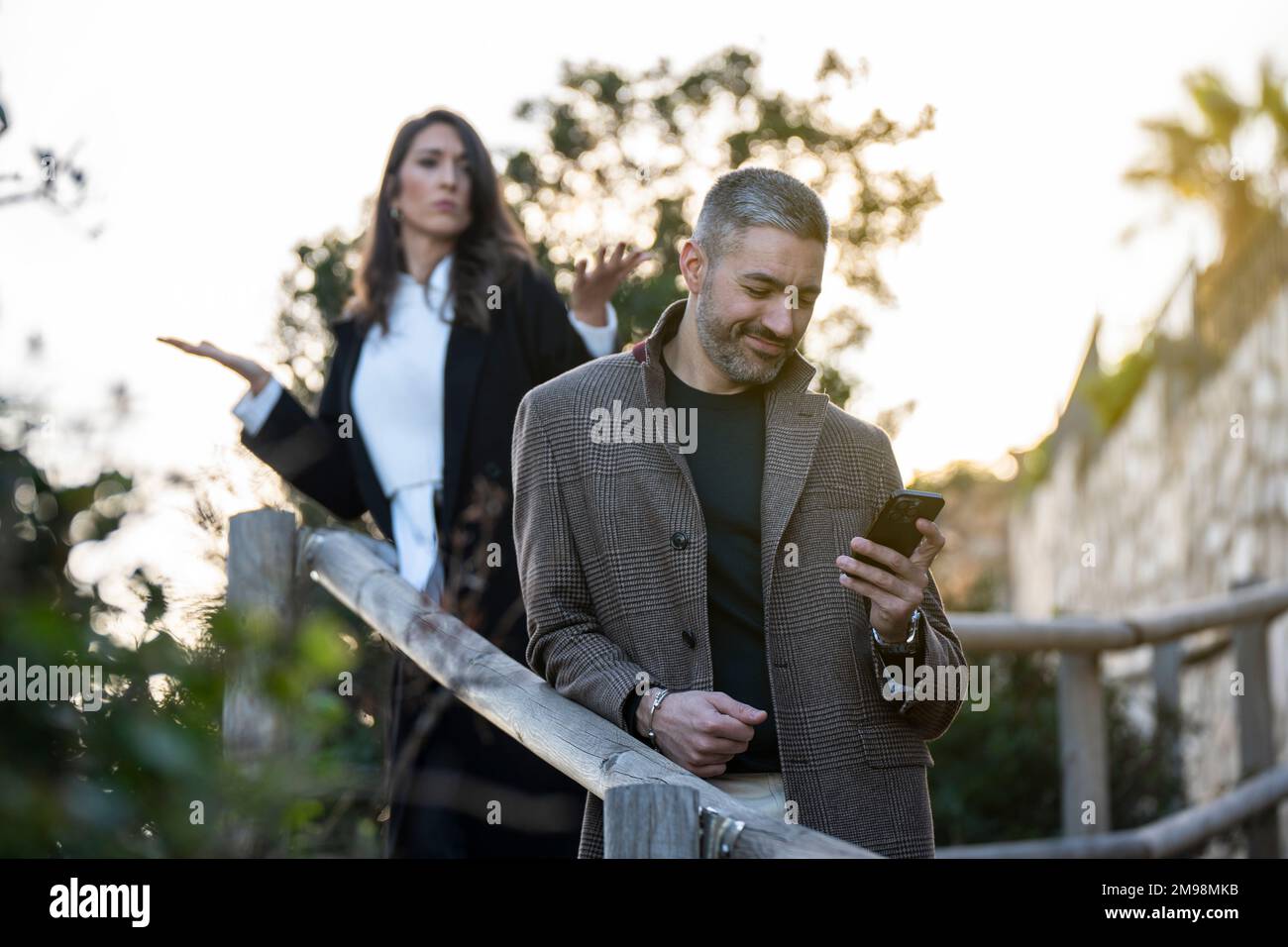 Ein Mann, der auf sein Handy schaut, mit einer wütenden Freundin dahinter Stockfoto