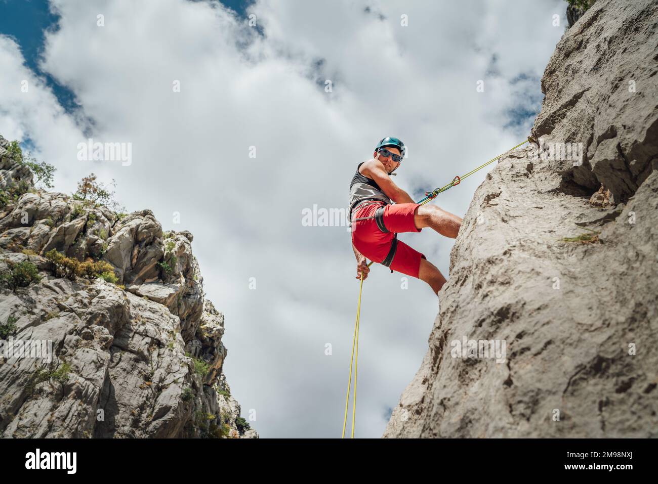 Aktiver Bergsteiger Mann mittleren Alters in Schutzhelm Blick auf Kamera, während Abseilen von Felswand mit Seil mit Sicherungsvorrichtung und Klettern ha Stockfoto