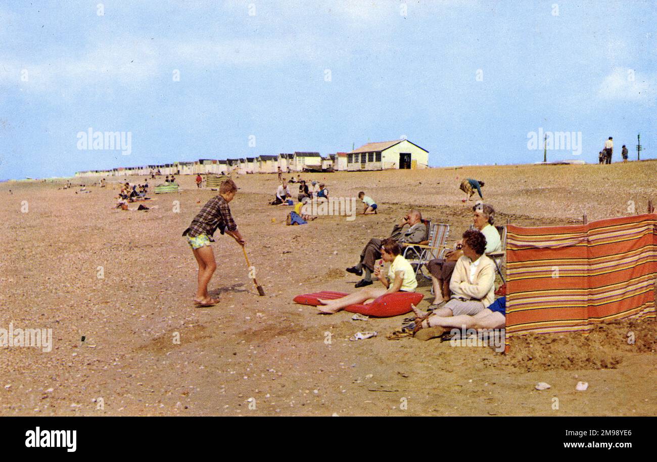 Holiday Beach Szene - Heacham, Norfolk - alle wesentlichen Merkmale des britischen Strandurlaubs sind hier - mehrere Schichten Kleidung, ein gestreifter Windschutz, Opa nickt in einem ausklappbaren Stuhl, ein hellroter Lolly, der widerwillig verzehrt wird, Schuhe, die getragen wurden, um dem Unbehagen beim Gehen auf Kieseln entgegenzuwirken, und eine Reihe von verschlossenen Strandhütten, die sich bis in die Unendlichkeit wiederholten Stockfoto