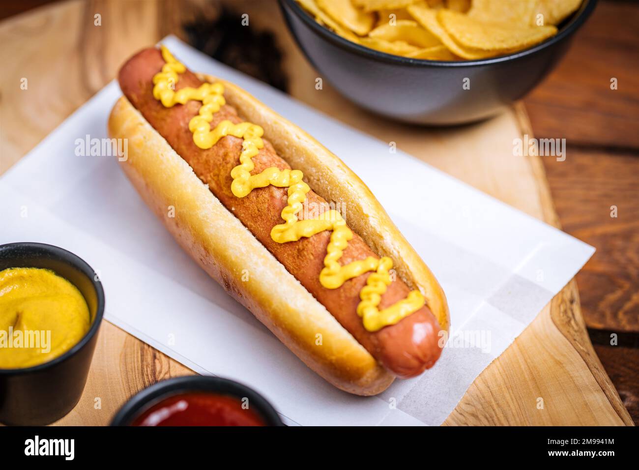 Klassischer Fast-Food-Hotdog mit Senf auf einem rustikalen Holzbrett. Serviert mit Kartoffelchips und Soßen. Stockfoto