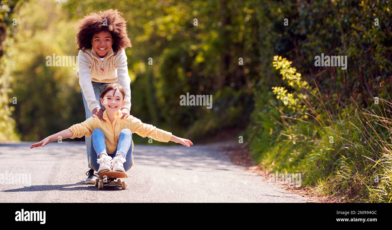 Kinder, Die Spaß Haben Mit Dem Jungen, Das Mädchen Auf Dem Skateboard Auf Der Country Road Schiebt Stockfoto