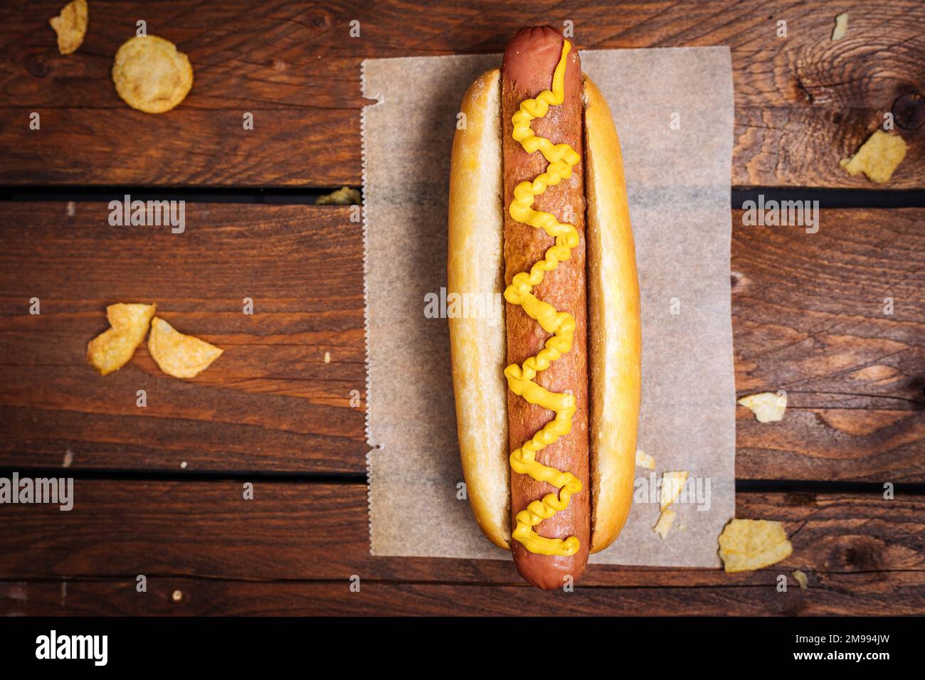 Klassischer Fast-Food-Hotdog mit Senf auf einem rustikalen Holzbrett. Serviert mit Kartoffelchips und Soßen. Stockfoto