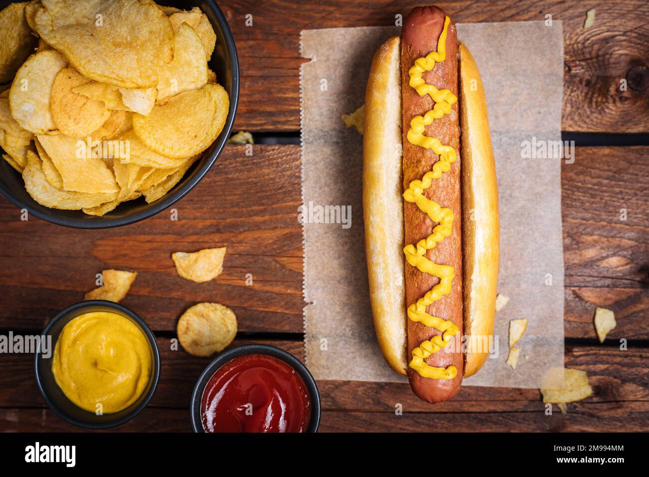 Klassischer Fast-Food-Hotdog mit Senf auf einem rustikalen Holzbrett. Serviert mit Kartoffelchips und Soßen. Stockfoto