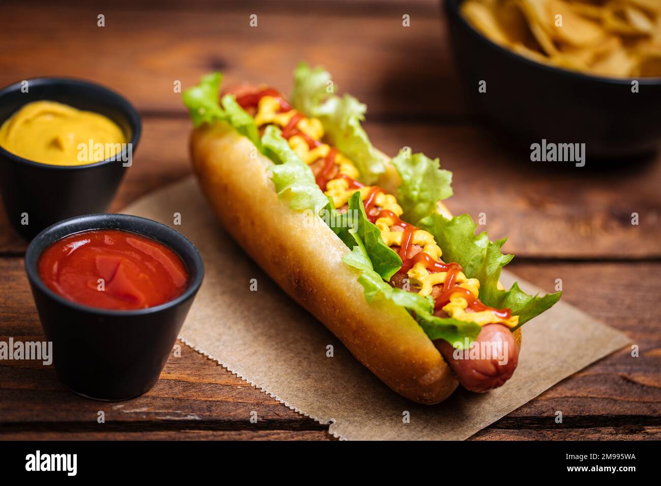 Hot Dog mit Senf, Ketchup und grünem Salat im Brötchen, serviert mit Kartoffelchips auf einem rustikalen Holzbrett. Fast Food und köstlich. Stockfoto