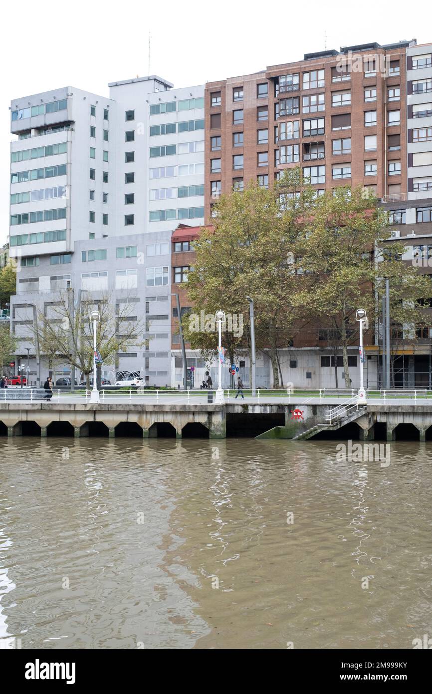 Blick über den Fluss Nervíon in Bilbao, Baskenland, mit Gebäuden dahinter und einer gefliesten Straßenkunst am Rand des Flusses, die zum Fluss führt Stockfoto