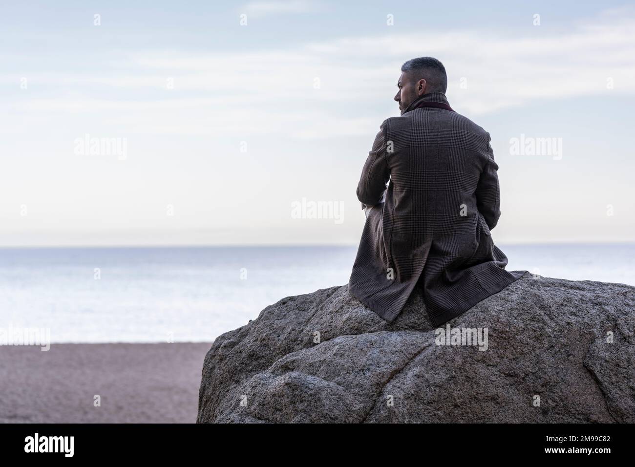 Ein Mann sitzt auf einem Felsen am Strand und schaut aufs Meer Stockfoto