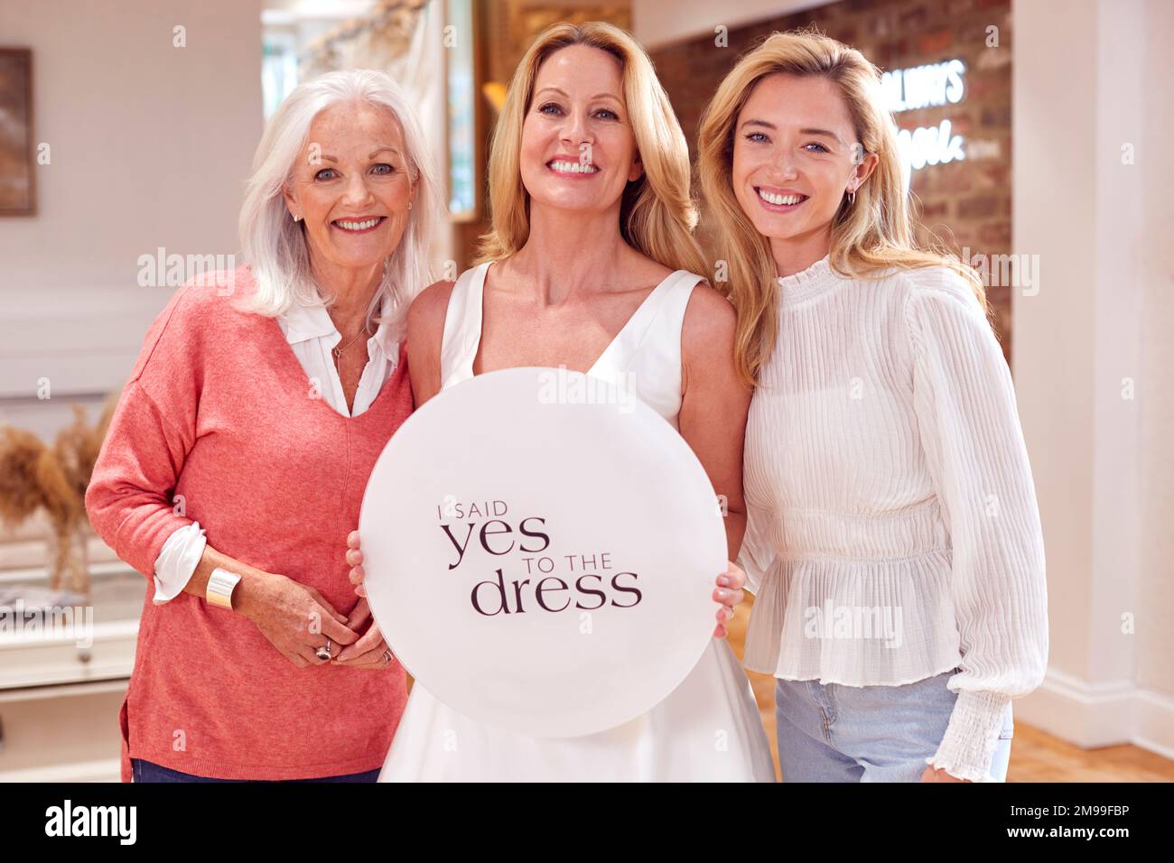 Großmutter Mit Erwachsener Tochter Und Enkelin Im Brautladen Mit „Ja Zum Kleid“-Schild Stockfoto