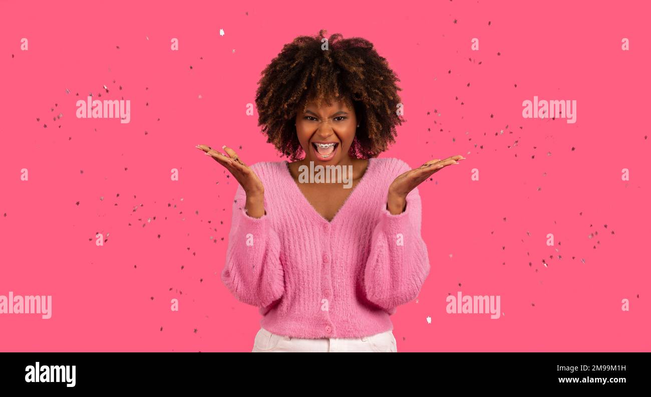 Fröhliche junge afroamerikanische lockige Frau mit einer Zahnspange mit offenem Mund wirft Glitzer und feiert Feiertage Stockfoto