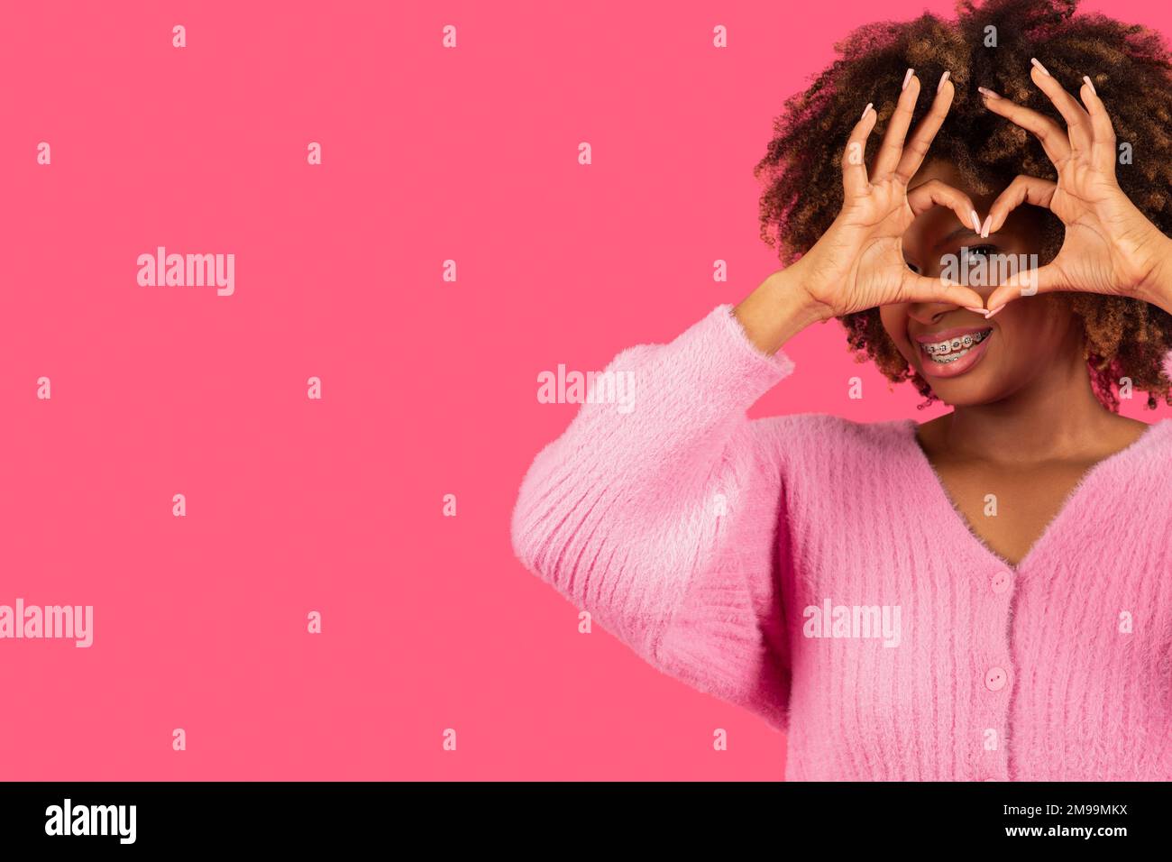 Lächelnde junge afroamerikanische, lockige Frau mit einer Zahnspange, die ein Herzzeichen zeigt, mit Händen in Augennähe, viel Spaß Stockfoto