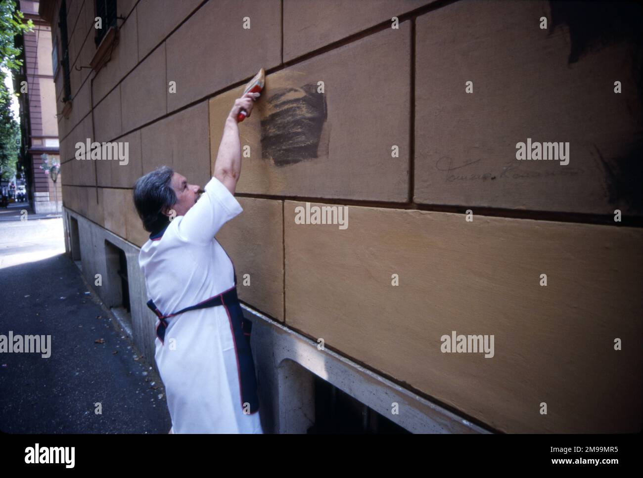 Eine Seniorin, eine Italienerin, legt eine Farbe an eine Wand, um Graffiti auf einem römischen Gebäude zu verdecken Stockfoto