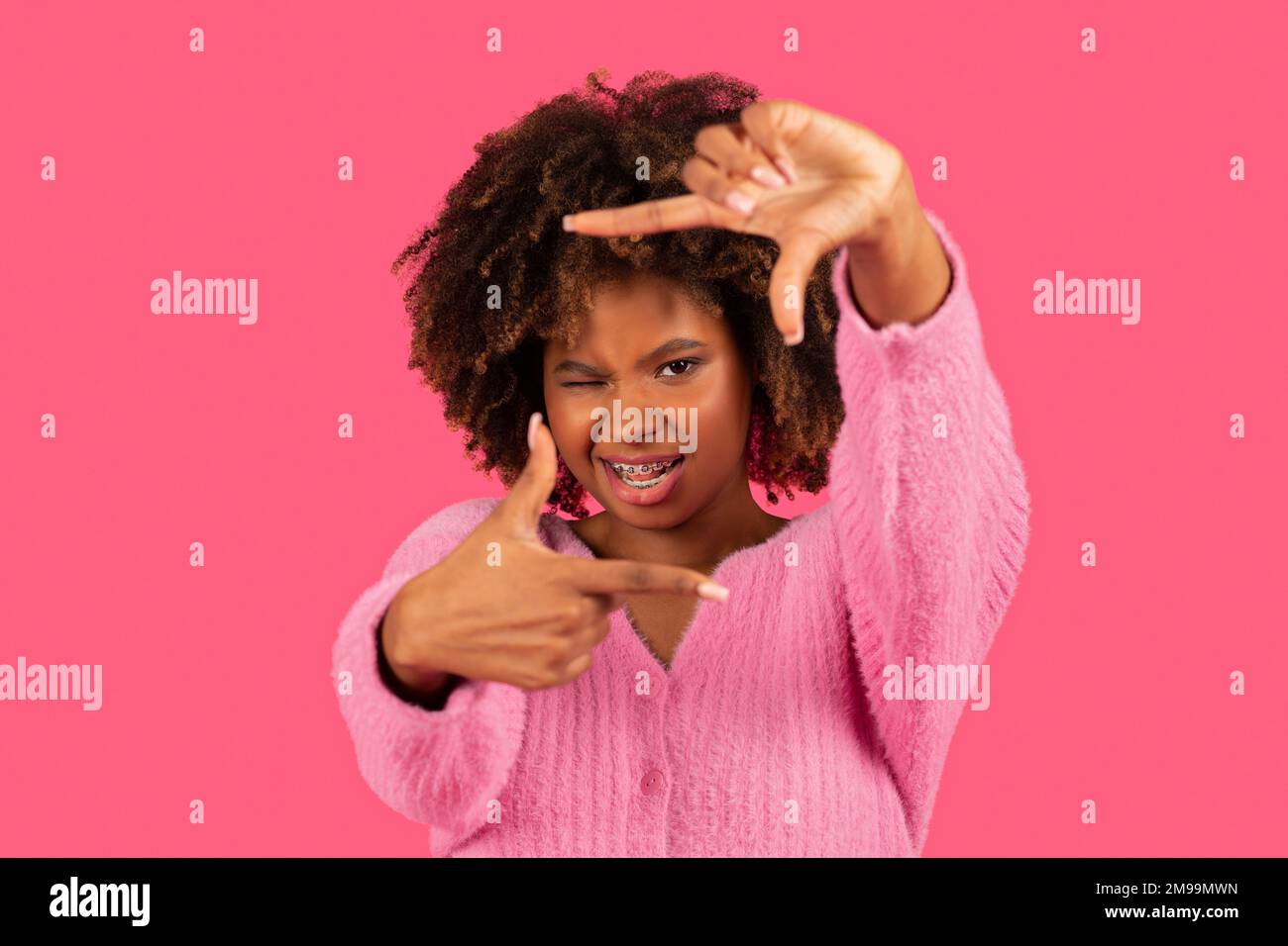 Fröhliche junge afroamerikanische lockige Frau mit Zahnspangen zeigt ein Schild, macht einen Rahmen mit Händen, schaut hindurch Stockfoto