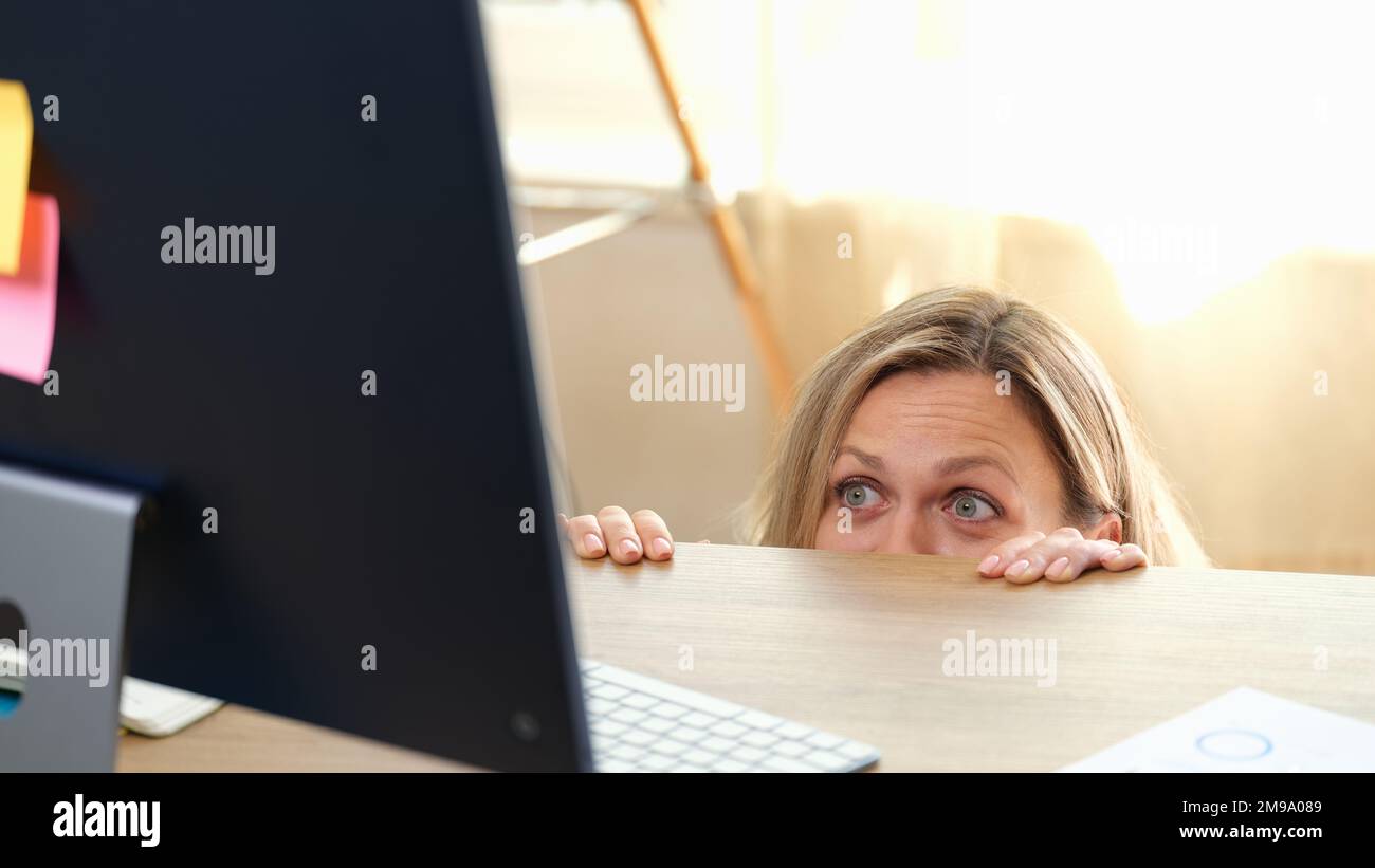 Frau versteckt sich hinter dem Schreibtisch und schaut auf den Computermonitor. Stockfoto