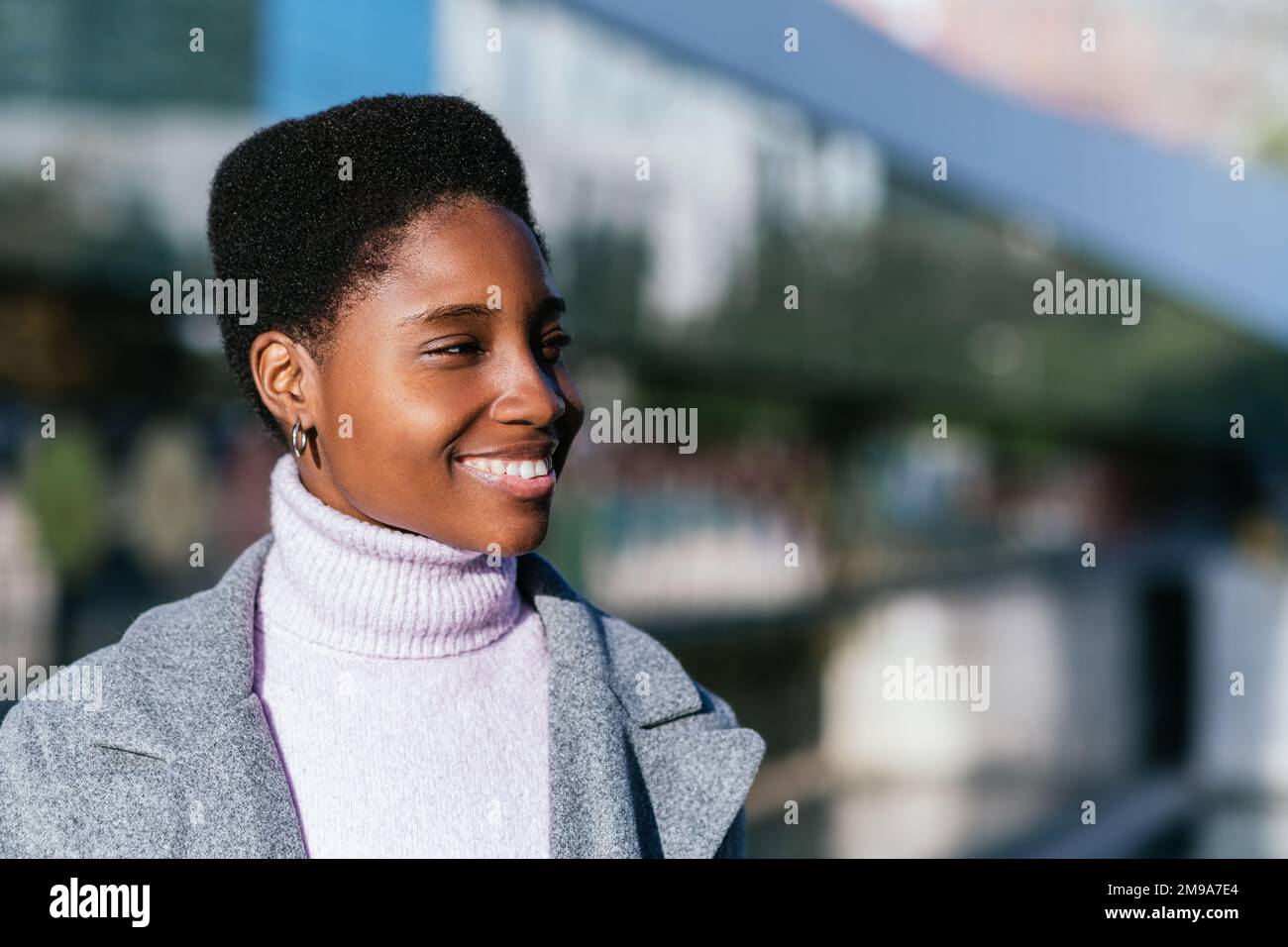 Positive schwarze Frau in stilvollem Mantel und Pullover mit kurzen Haaren, die lächeln und am Tag auf die Straße der Stadt blicken Stockfoto