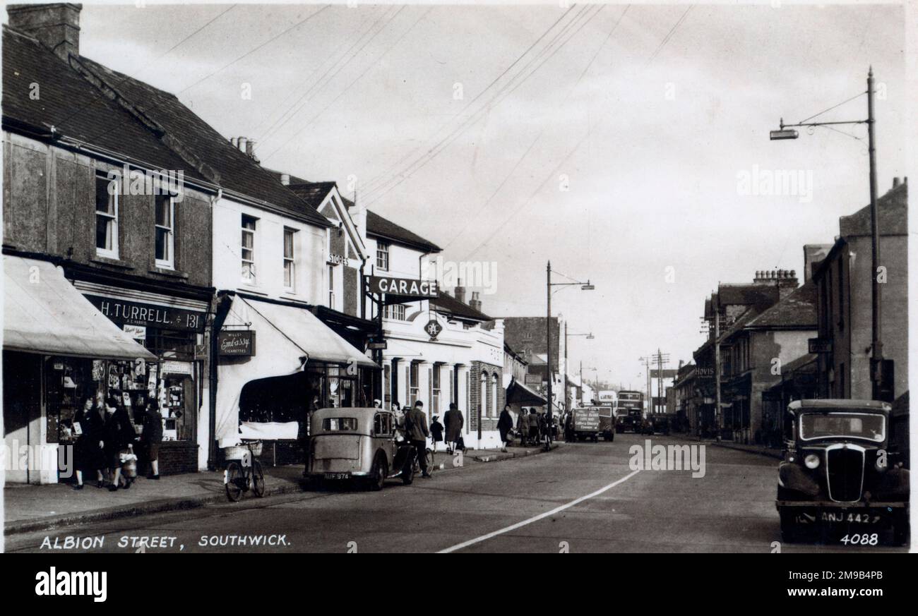 Das Auto auf der rechten Seite hat das Nummernschild ANJ442. Die Aussicht zeigt Turrells Stores mit einer Anzeige für Embassy Zigaretten und einer Garage dahinter. Der Bus ist in der Ferne. Stockfoto
