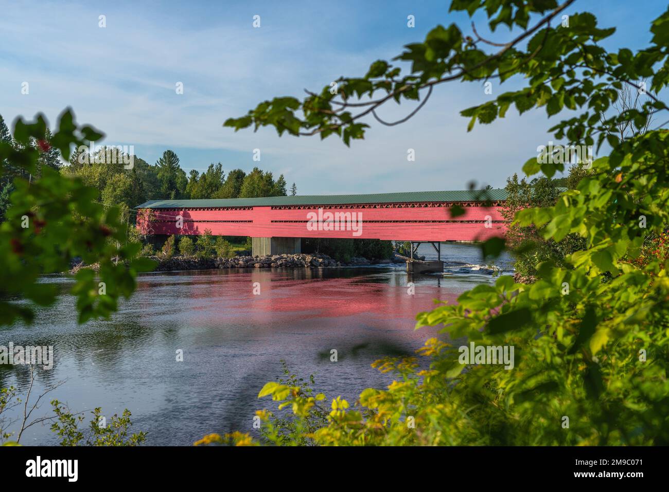 Ein gerahmter Blick auf eine rote, hölzerne überdachte Brücke über einen großen Fluss mit dem borealen Wald um ihn herum, Laurentianer, Quebec, Kanada. In einem sonnigen Sommer aufgenommen Stockfoto