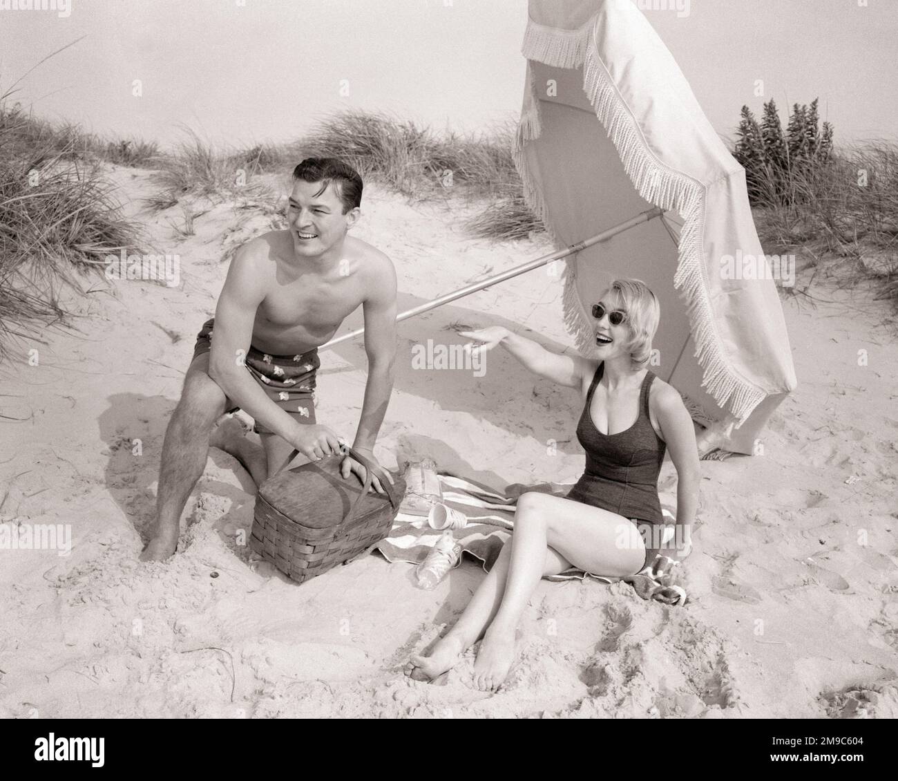 1960S PAAR AM STRAND IN BADEANZÜGEN MANN, DER GERADE EINEN PICKNICKKORB ÖFFNET FRAU, DIE LÄCHELT UND AUF LINKS IN DER SONNE ZEIGT REGENSCHIRM - B12268 DEB001 HARS HEALINESS COPY SPACE FRIENDSHIP DURCHGEHENDE HALBE LÄNGE SUNNY SHADE B&W ZEIT OFF SHORE GLÜCK FRÖHLICHE ENTDECKUNG URLAUBSREISE AUFREGUNG NIEDRIGE ERHOLUNGSSTRÄNDE URLAUB IM FREIEN FREUDENFREUDIGES PICKNICK SANDY STILVOLLE DEB001 ZUSAMMENARBEIT LIESS ENTSPANNUNG ZUSAMMENWESENHEITSURLAUBE JUNGER ERWACHSENER MANN JUNGE ERWACHSENE FRAU SCHWARZ-WEISS WEISSE ETHNISCHE HERKUNFT ALTMODISCH Stockfoto