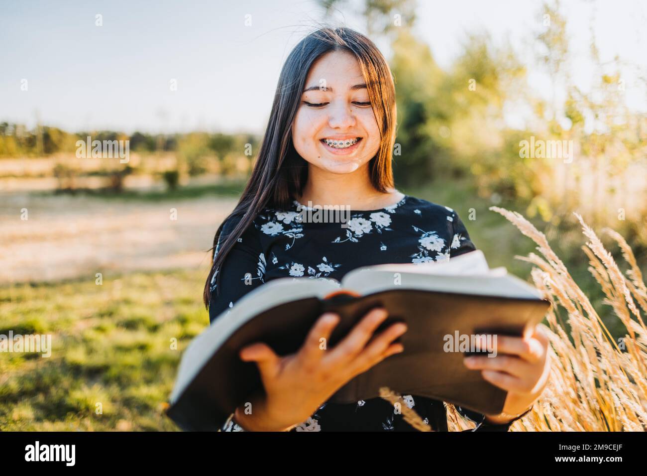 Unschuldiges, junges, religiöses Mädchen mit Zahnspangen, das die bibel studiert, draußen auf dem Feld bei Sonnenuntergang. Spirituelle Wiederbelebung Stockfoto