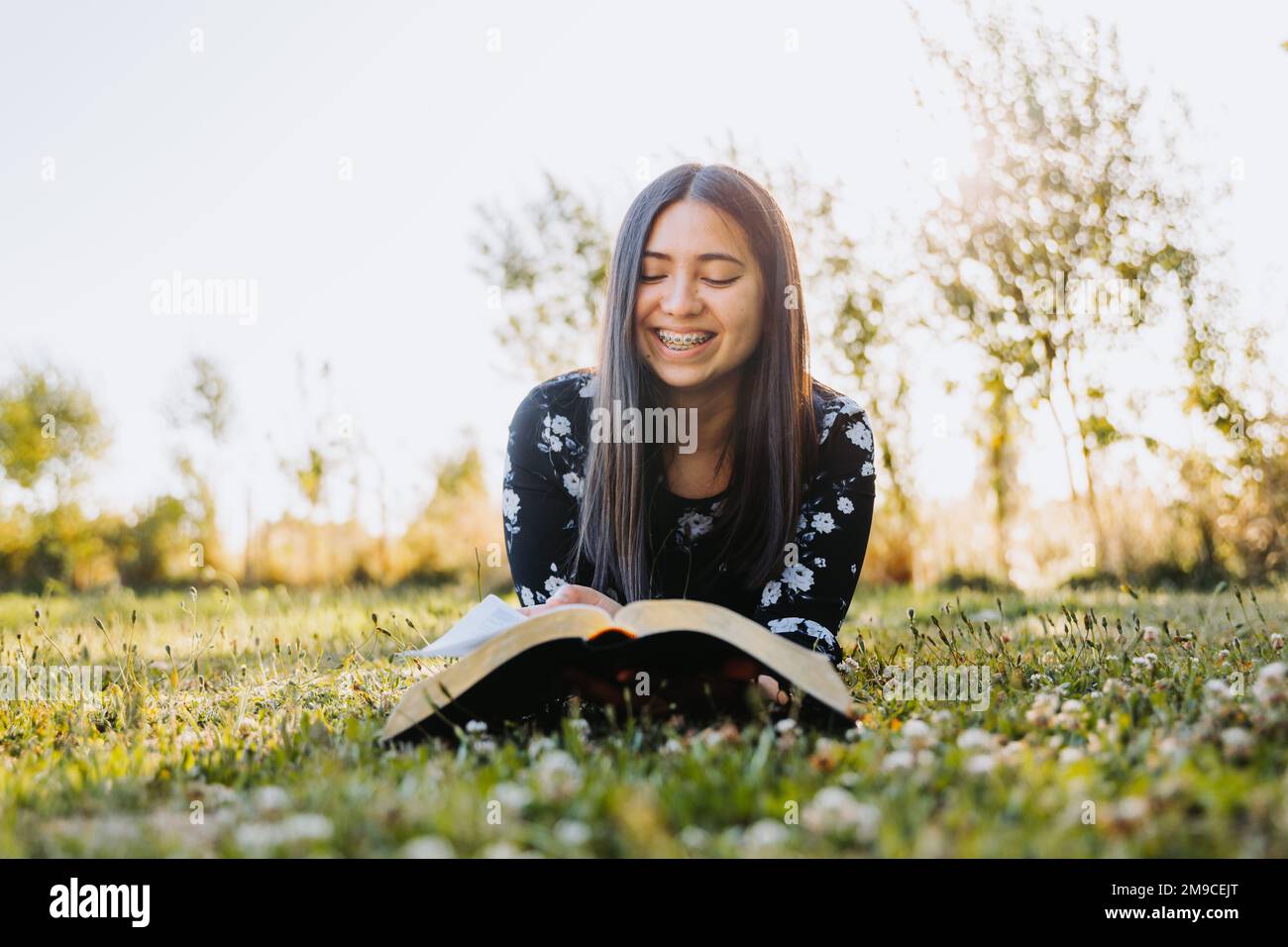 Junge lächelnde Frau mit Zahnspange, Gläubige, die ihre bibel studiert, während sie auf dem Gras liegt, in einem natürlichen Raum. Stockfoto