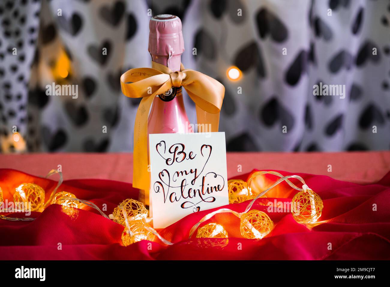Valentinstag Dekorationsidee, rosafarbener Champagner mit goldenem Band und Notiz auf einem romantischen rosafarbenen Tischtuch in Schwarz und Weiß mit herzförmigem Hintergrund Stockfoto