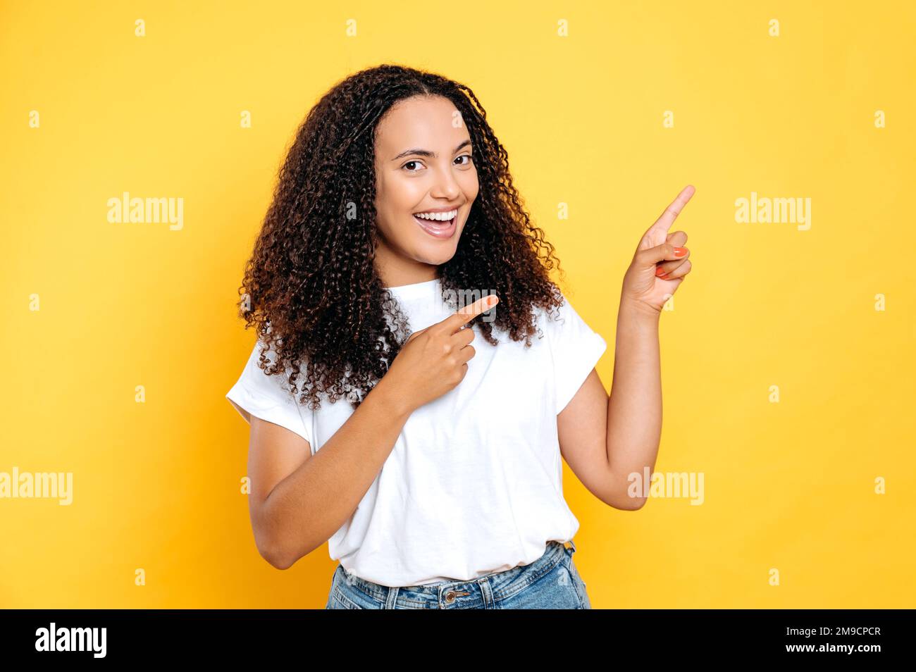 Fröhliche, gemischte Rasse Frau mit lockigem Haar, trägt ein weißes T-Shirt, zeigt mit beiden Händen auf einen leeren Raum, schaut in die Kamera, lächelt, steht über isoliertem gelben Hintergrund Stockfoto