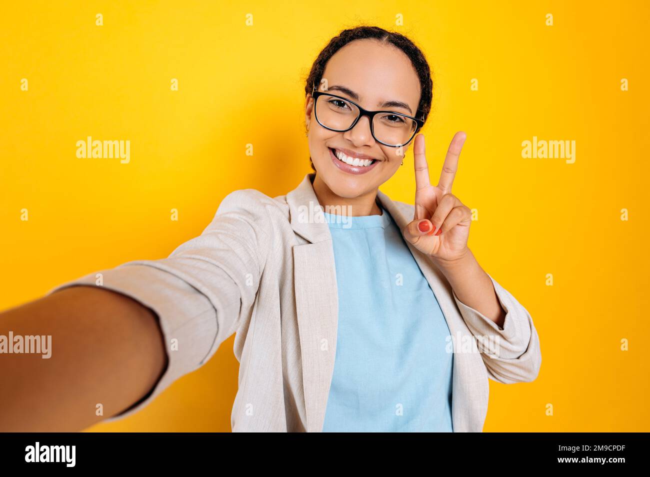 Hübsche, positiv charismatische hispanische oder brasilianische Frau mit Brille, macht ein Selfie-Foto auf ihrem Smartphone, schaut in die Kamera, lächelt, zeigt Frieden mit ihren Fingern, gelber isolierter Hintergrund Stockfoto