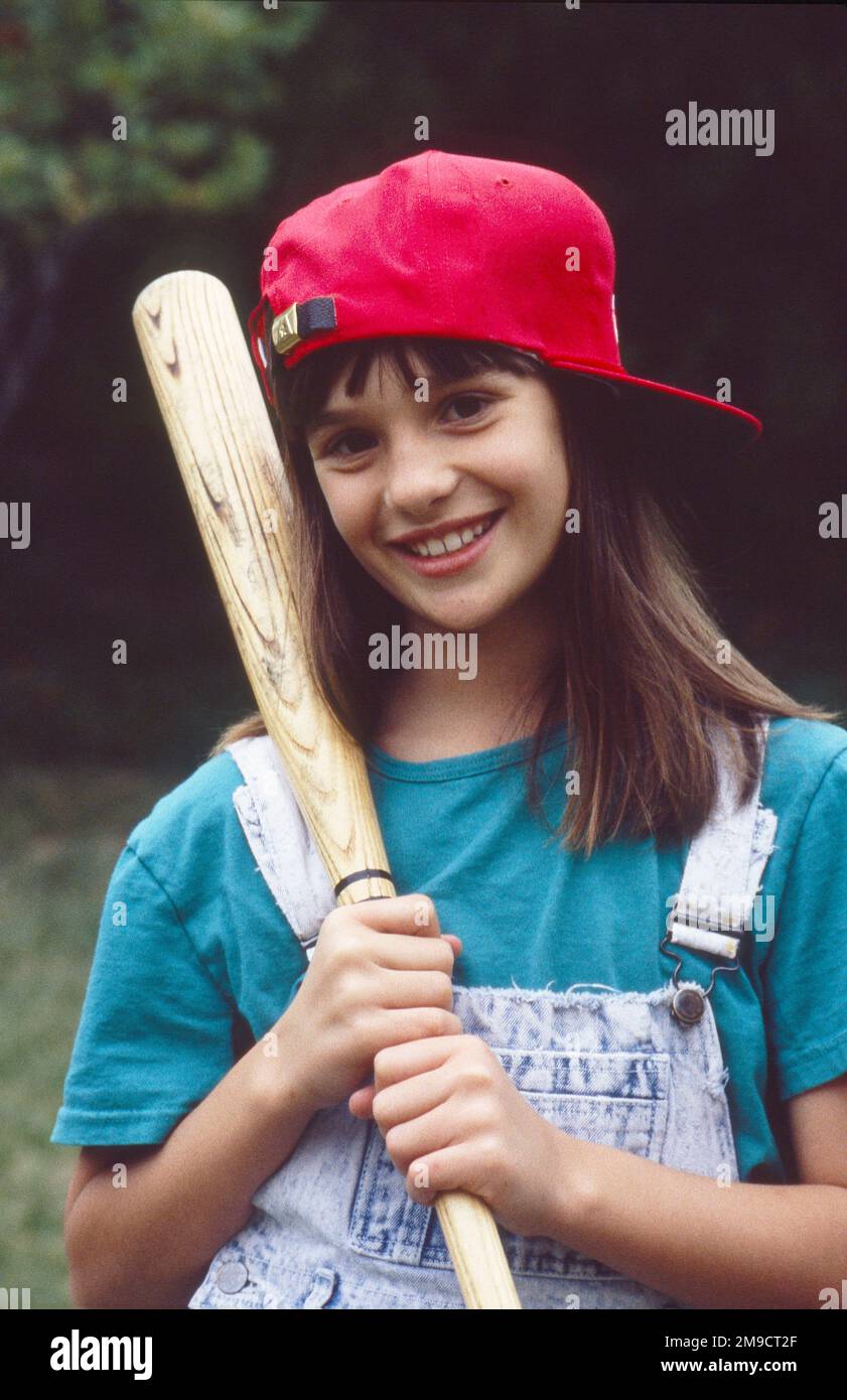 Ein kleines Tomboy-Mädchen, das mit einer Baseballkappe auf dem Rücken posiert, einen Baseballschläger hält und in die Kamera lächelt Stockfoto