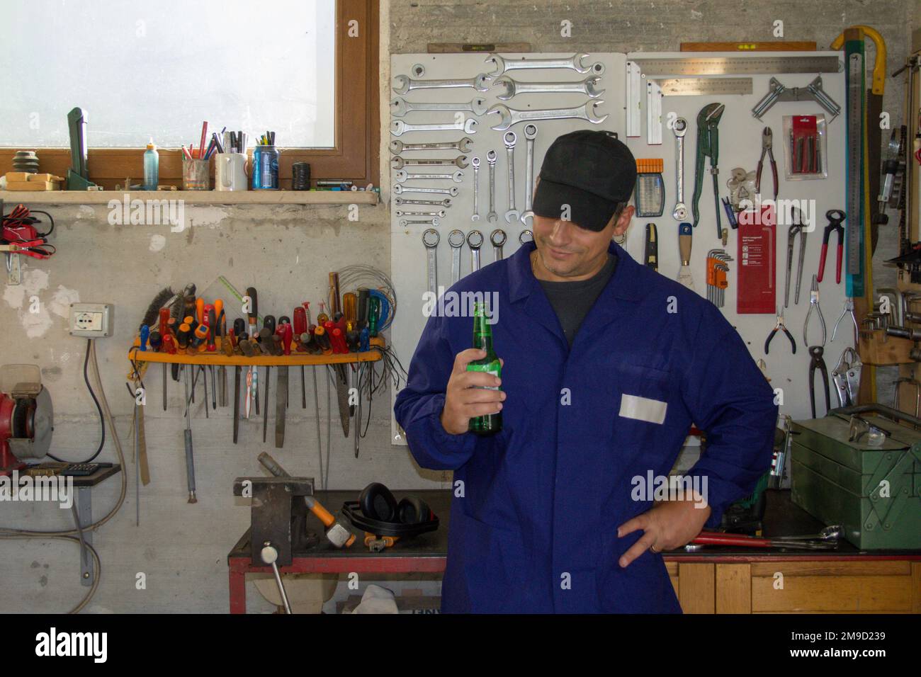 Bild eines Mannes im Overall mit einer Flasche Bier in seiner Werkstatt. Pause, entspannen Sie sich nach der Arbeit. Stockfoto